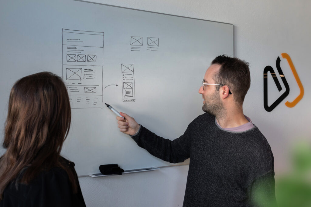 Twee mensen staan voor een whiteboard met een schets van een website lay-out. Bernard wijst met een stift naar verschillende elementen op de schets, terwijl hij uitleg geeft aan de persoon naast hem. Op de achtergrond is een deel van het Webreact-logo zichtbaar op de muur.