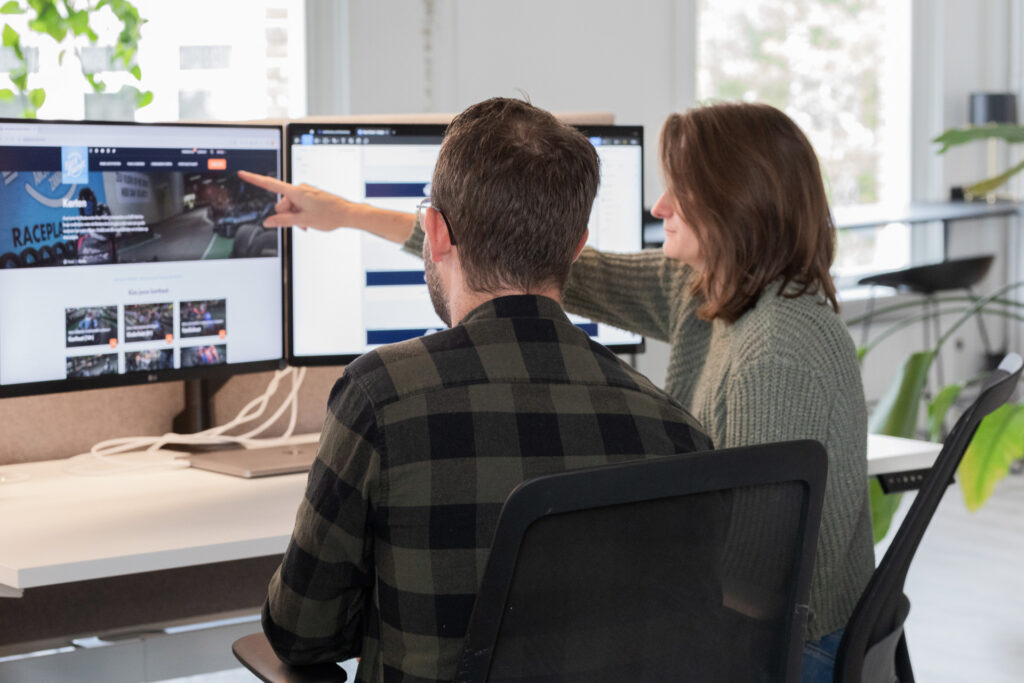 Twee personen zitten achter een bureau en kijken naar een computerscherm waarop een website wordt weergegeven. De vrouw wijst iets aan op het scherm terwijl de man aandachtig kijkt. Er staan meerdere monitors op het bureau, en de ruimte is licht en modern ingericht met enkele planten op de achtergrond.